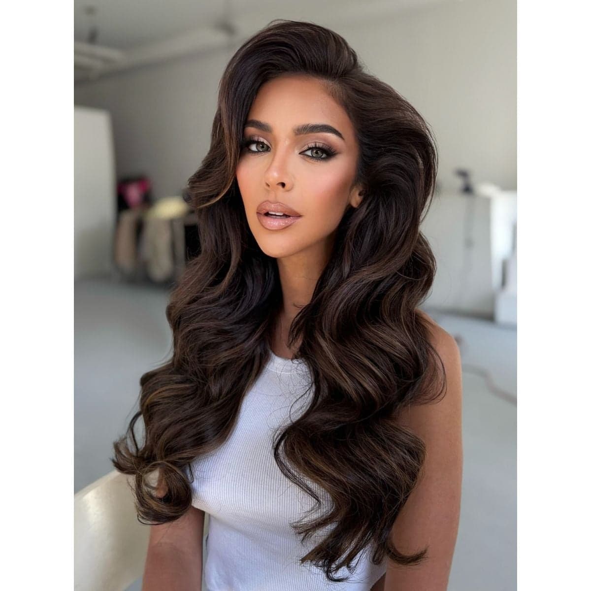 A woman with long, voluminous dark brunette hair styled in glamorous, soft waves. She’s wearing a white sleeveless top and posing in natural light, showcasing glossy, multidimensional color and full-bodied texture.